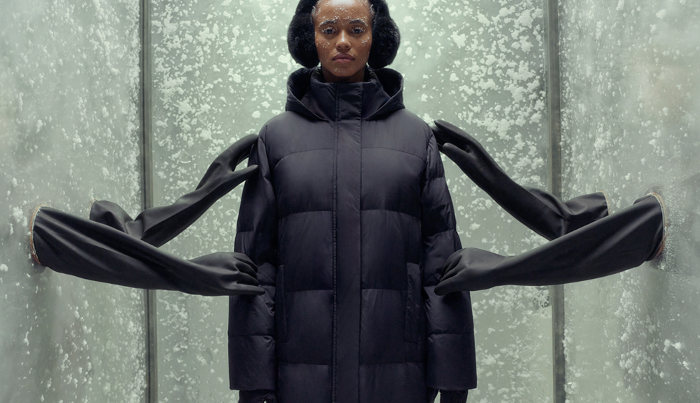 Cadru mediu cu o femeie care pozează într-o haină lungă neagră matlasată, cu glugă. Poartă și căști de urechi pufoase și mănuși negre.